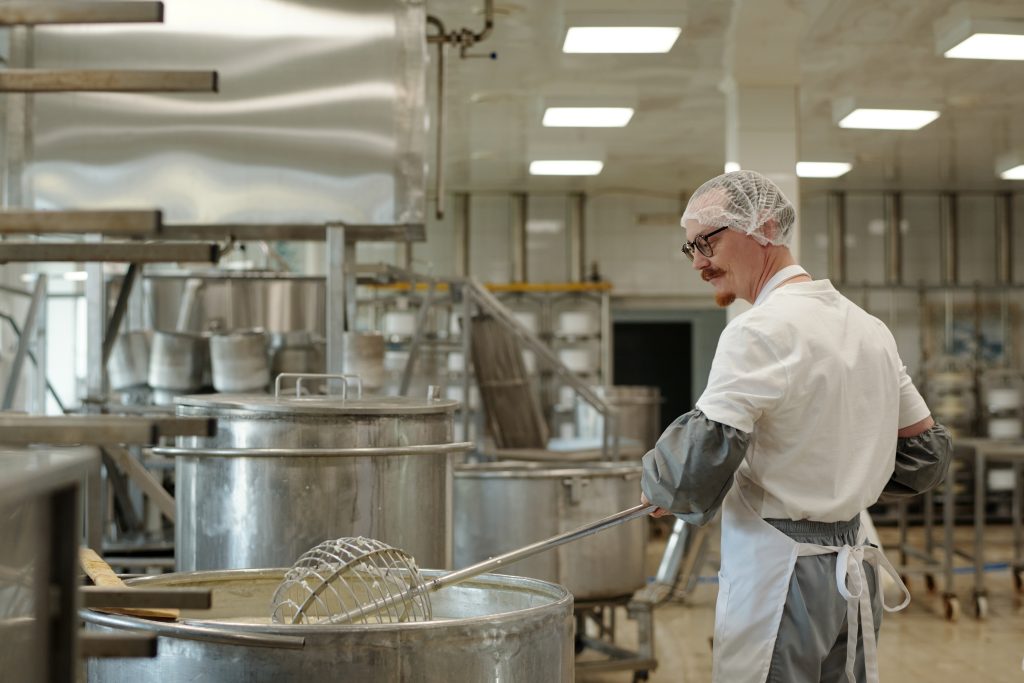 Chef preparing food in large commercial kitchen, surrounded by industrial cooking pots and machinery. Wearing protective gear and involved in food preparation process.